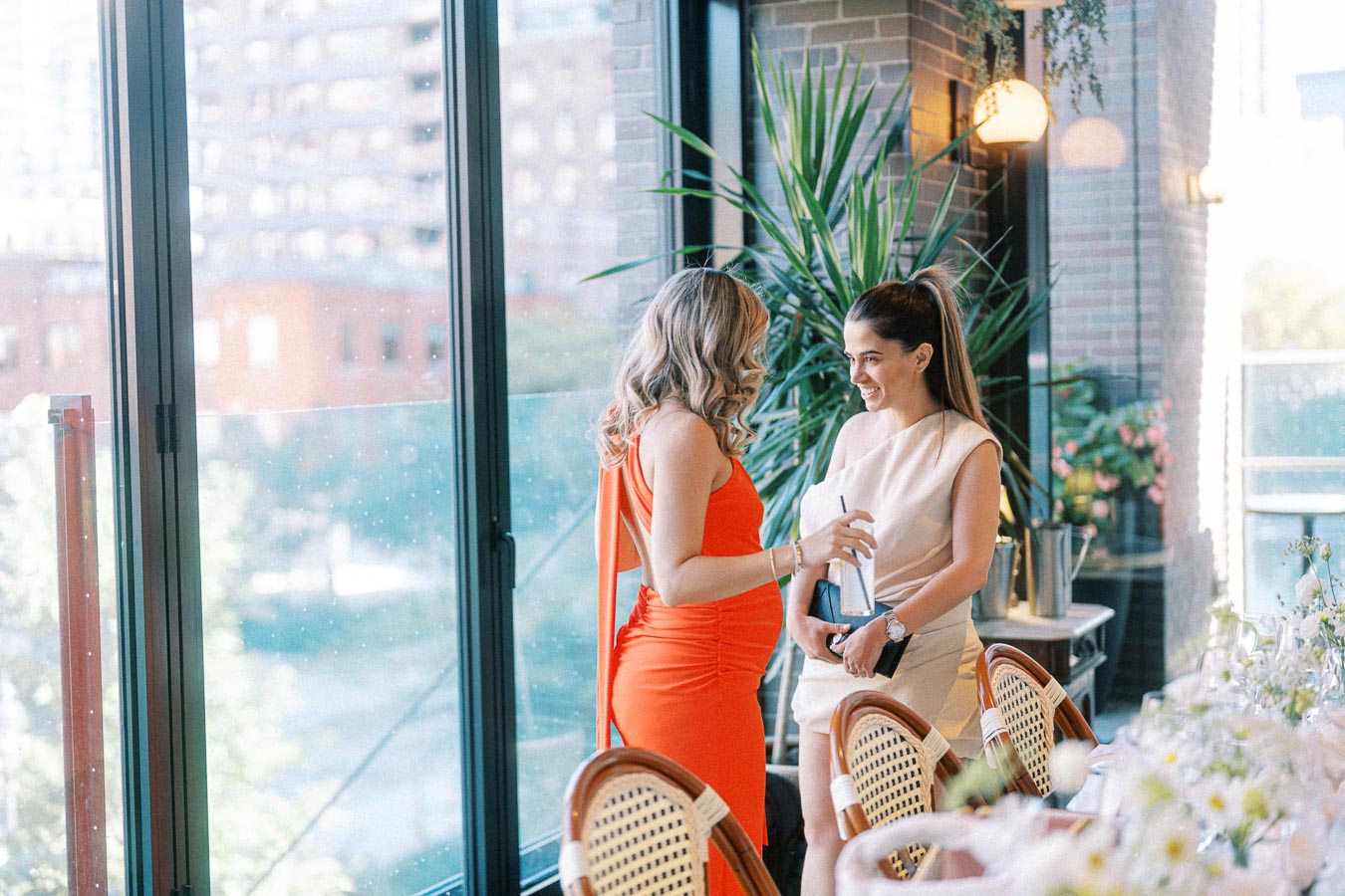 Two women in elegant dresses engaging in conversation at a beautifully set dining table by a large window with a city view, highlighting a sophisticated event or gathering in an urban setting.