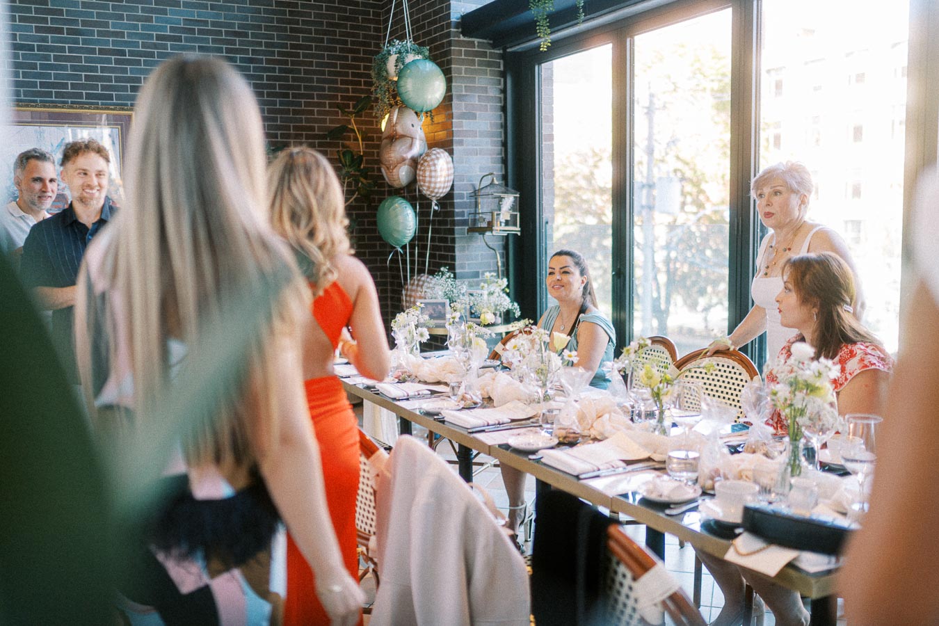 A lively gathering of people at a beautifully decorated indoor event space with a long table set for a celebration. Balloons and flowers adorn the setting, as guests engage in conversation and enjoy the sunlight streaming through large windows.