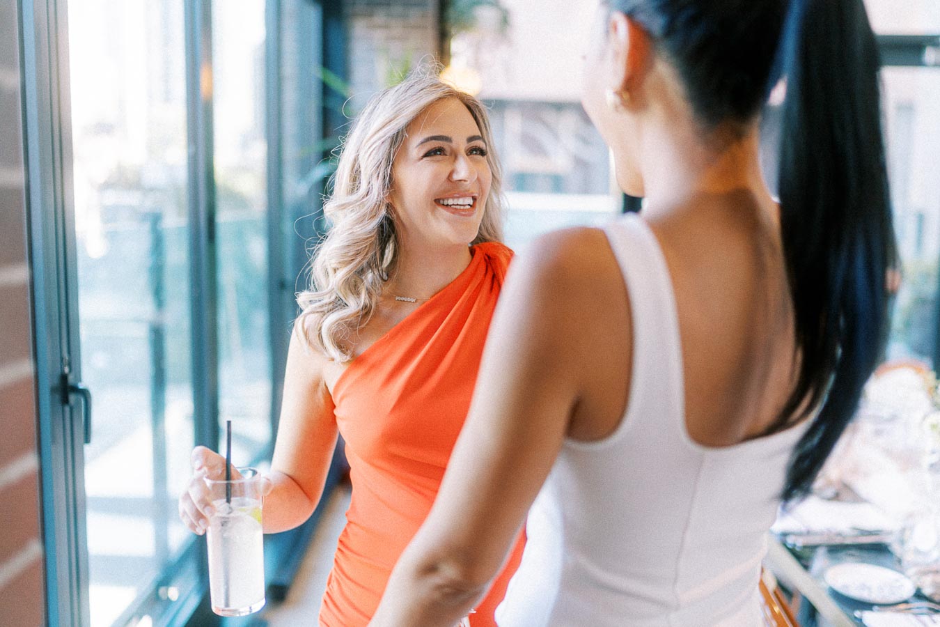 Two women smiling and engaging in a friendly conversation by a window, one wearing a vibrant orange dress and holding a glass with a drink. Bright indoor setting with natural light.