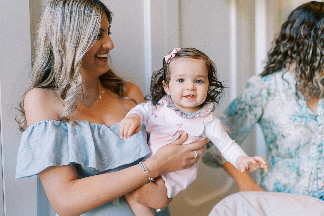 A smiling woman holds a happy baby wearing a pink outfit with floral details, both enjoying a joyful moment indoors.