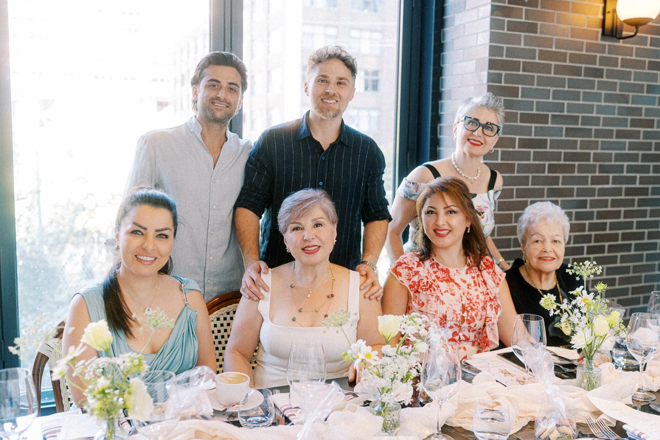 A joyful family gathering around a dining table set for celebration, with flowers, dishes, and smiles. The group includes older and younger adults enjoying a meal in a bright room with large windows and a brick wall.
