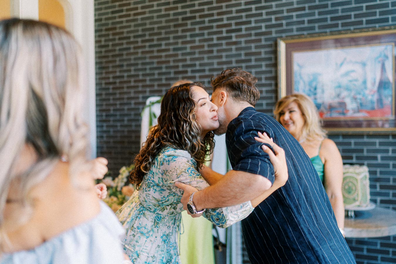 Friends greeting each other with a kiss on the cheek at a social gathering, with people smiling in the background, creating a warm and welcoming atmosphere.