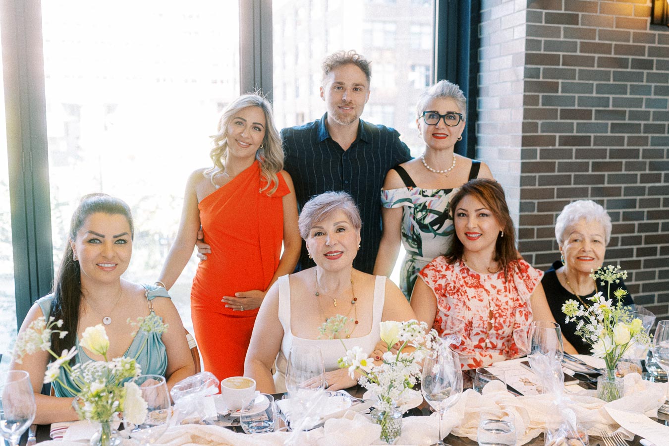 A group of seven people smiling at a restaurant table, featuring a pregnant woman in an orange dress, surrounded by floral arrangements and tableware, set against a window with a cityscape view.