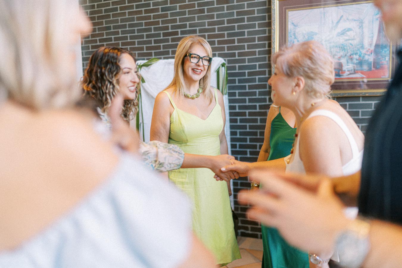 Group of women in colorful dresses engaging in a handshake at a social gathering, with a brick wall background.