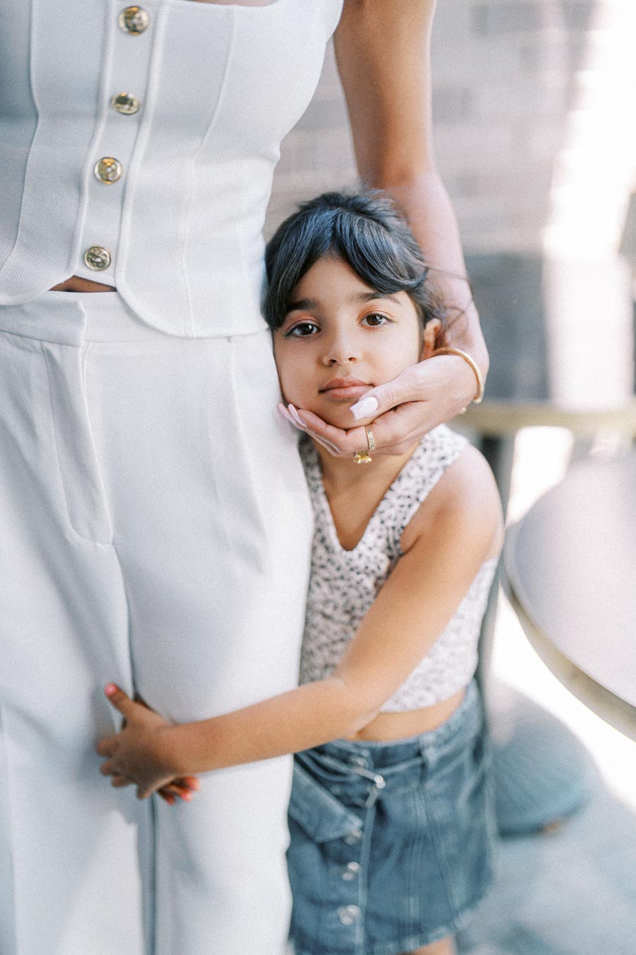 A young girl with dark hair hugging an adult wearing a white outfit, displaying a tender and affectionate moment.