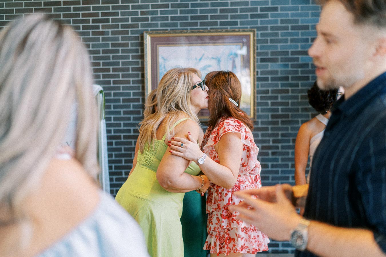 Women greeting each other with a kiss on the cheek at a social event, surrounded by blurred guests, against a brick wall background.