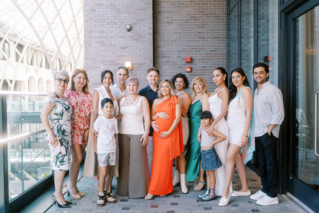 A diverse group of fourteen people, including adults and children, posing together for a family gathering in a modern, glass-enclosed building. The group is dressed in bright and elegant attire, with a pregnant woman in an orange dress at the center, creating a joyful and celebratory atmosphere.