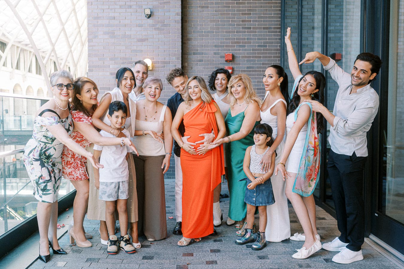 Group of people gathered indoors around a smiling pregnant woman, celebrating together, with outfits ranging from casual to semi-formal.
