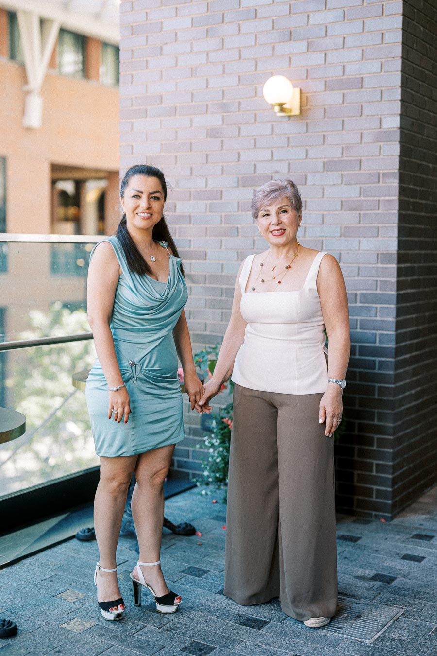 Two women holding hands and smiling at the camera, standing outdoors against a brick wall.