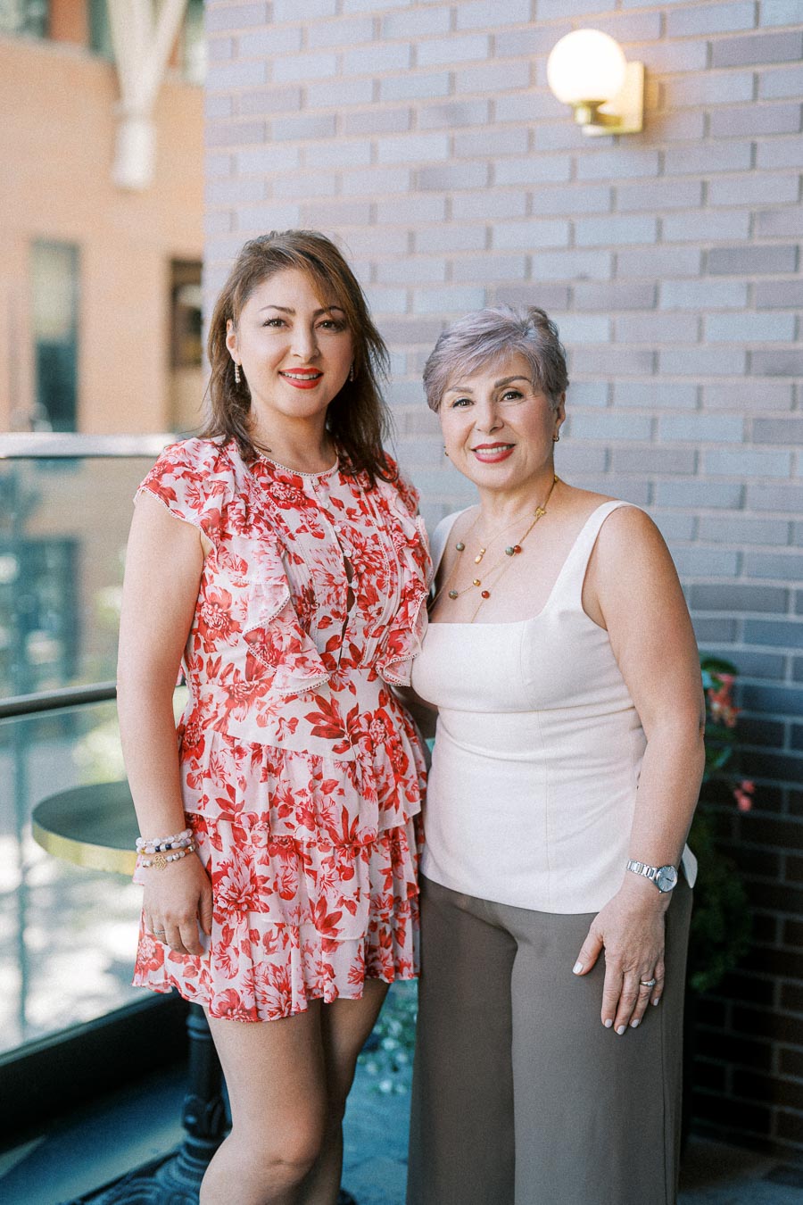 Two women smiling and posing together in front of a brick wall; one is wearing a red and white floral dress, and the other is dressed in a white top and brown pants. They appear to be enjoying a sunny day outdoors.