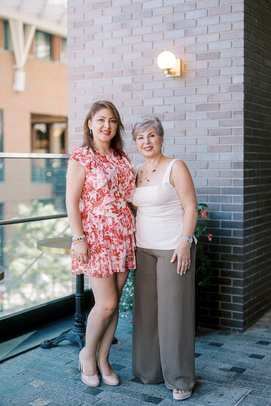 Two women smiling and posing together on an outdoor patio with a brick wall background, one in a floral dress and the other in a white top and brown pants.
