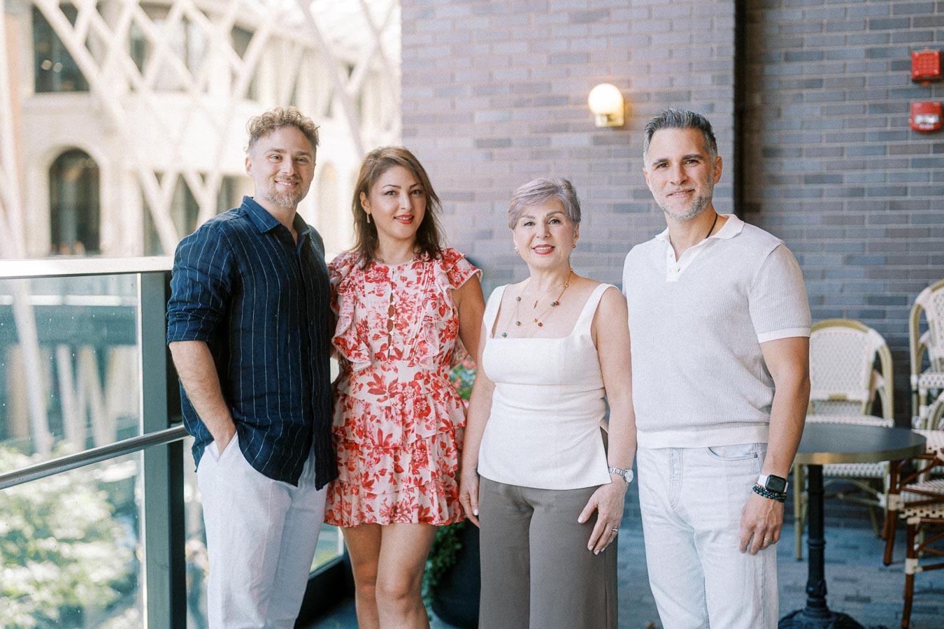 Four people posing together on a balcony with a modern architectural background, dressed in smart casual attire, smiling and enjoying a sunny day.