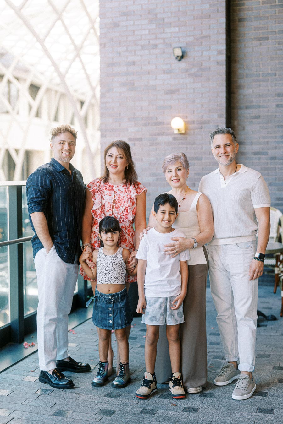Family smiling on outdoor patio against brick wall backdrop. Two adults and one senior stand with two children dressed casually, enjoying a sunny day. Modern architecture visible in the background