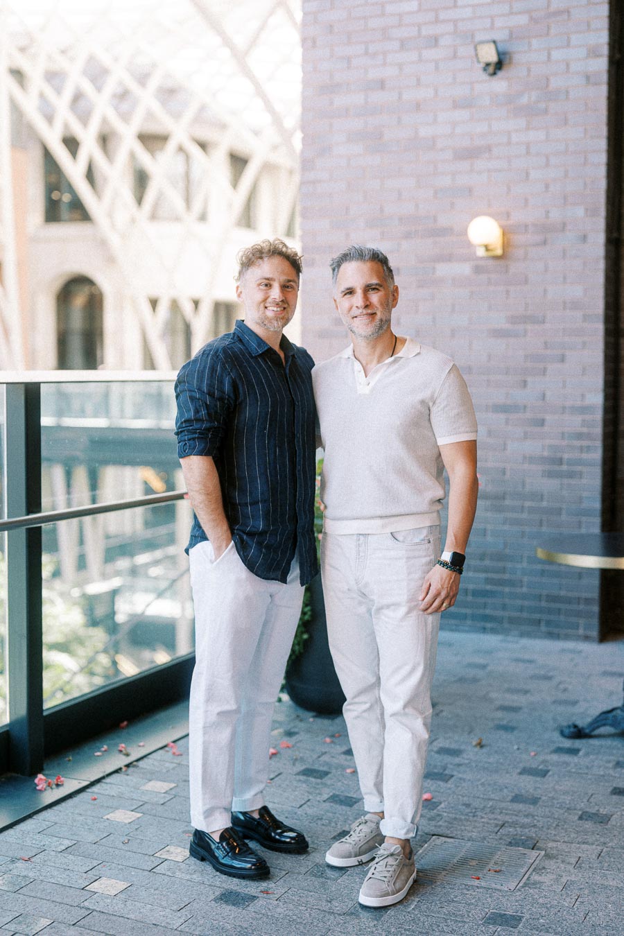Two men standing side by side outdoors wearing casual attire, one in a striped dark shirt and white pants, the other in a light polo and white pants, with a modern architectural building in the background.