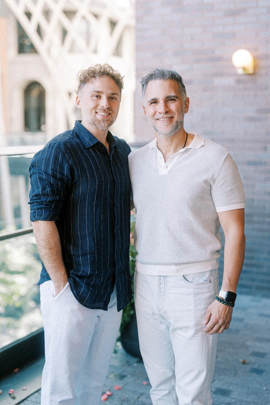 Two people smiling and standing together in a casual setting outside a building with a geometric architectural background, wearing stylish casual outfits.