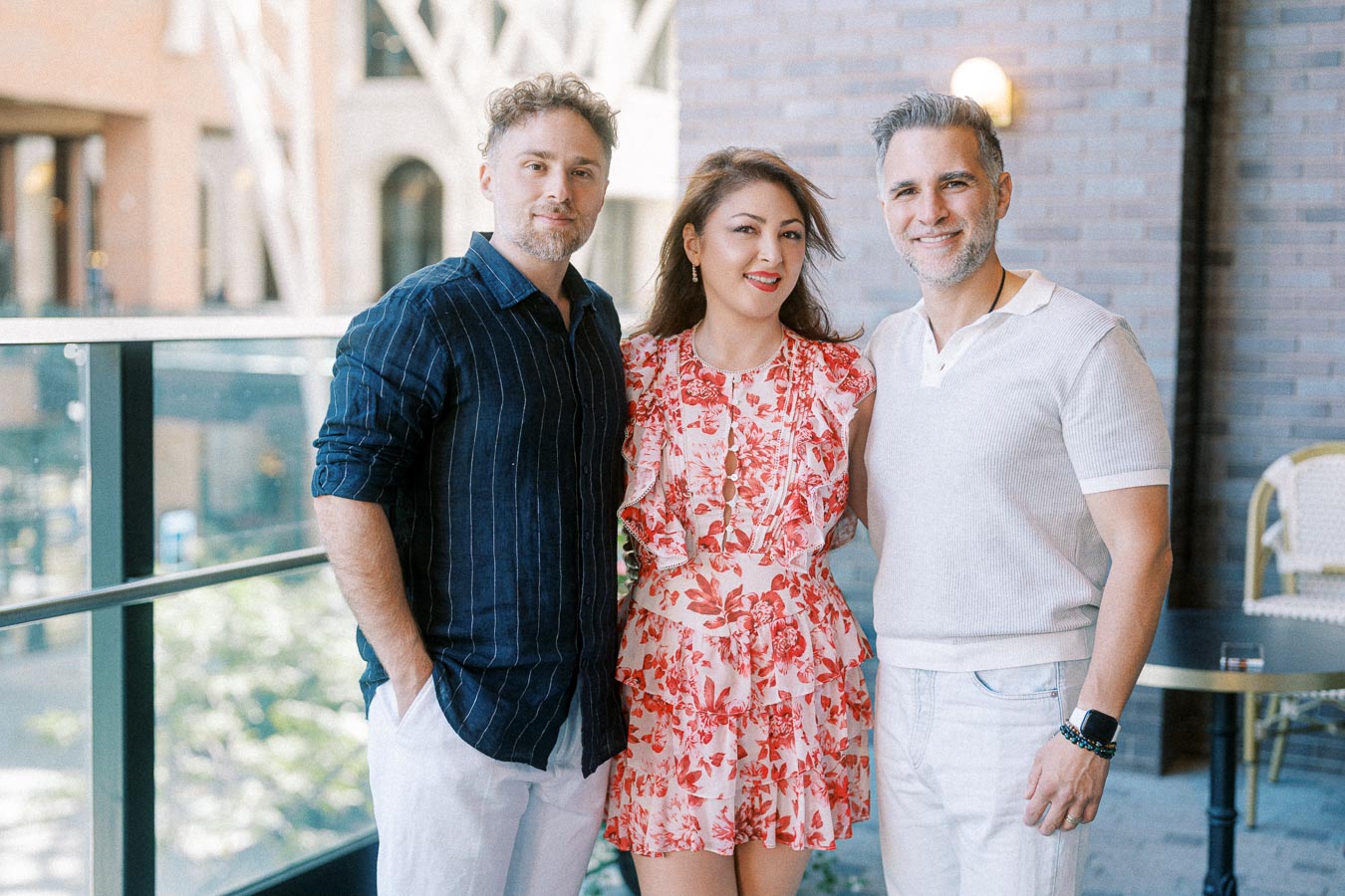A group of three people smiling and posing on a balcony, featuring a man in a dark shirt, a woman in a floral dress, and another man in a light shirt.