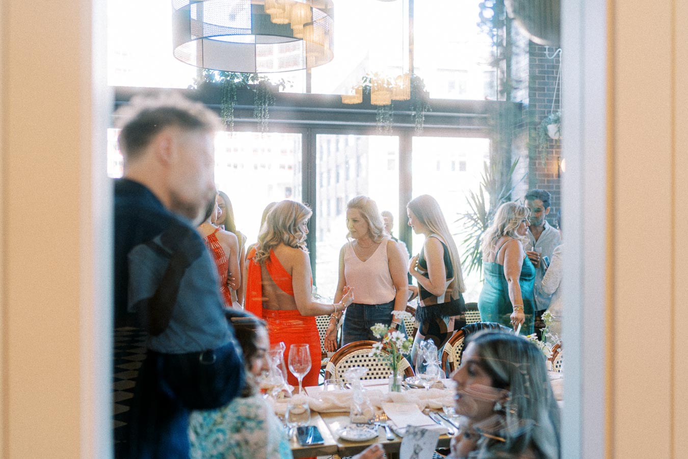 A group of people socializing at a stylish indoor event with large windows, decorated tables, and modern lighting fixtures.