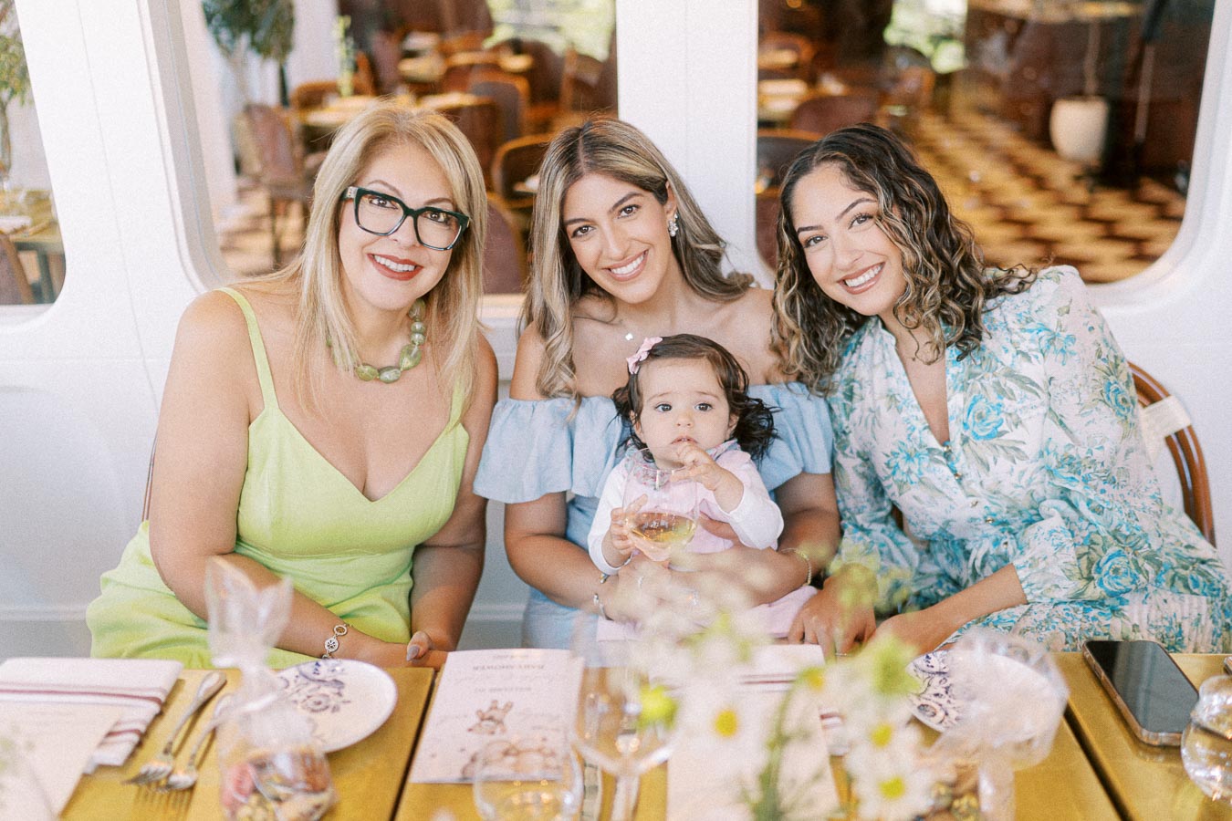 A joyful family gathering at a restaurant, featuring three women and a baby seated at a table decorated with floral arrangements and tableware. The women are smiling warmly at the camera, creating a lively and welcoming atmosphere.