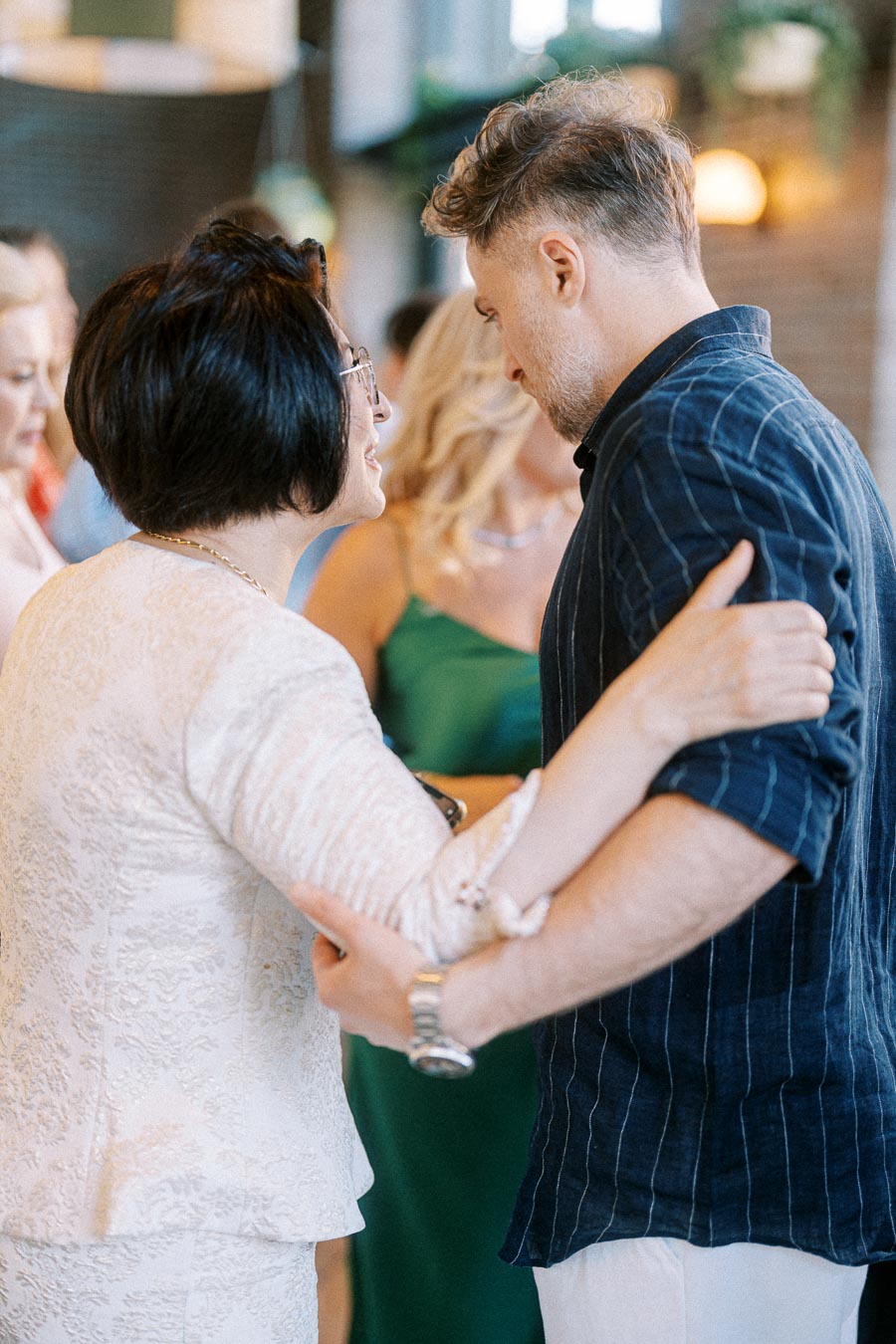 Two people in a heartfelt conversation at a social gathering, with one wearing a white lace outfit and the other in a blue striped shirt.