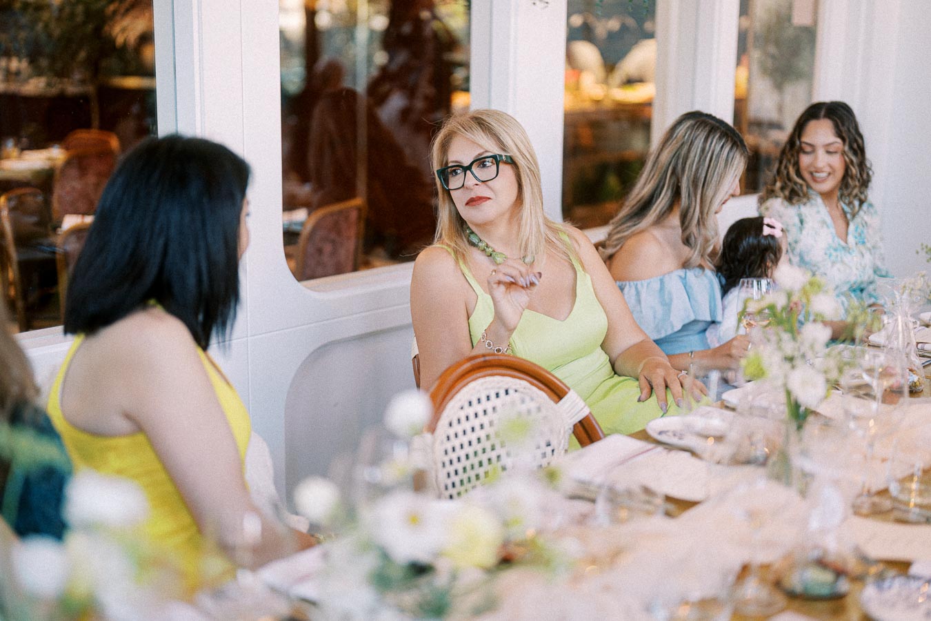 Group of women enjoying a lively conversation around a dining table at a cozy restaurant, surrounded by elegant decor and floral arrangements.