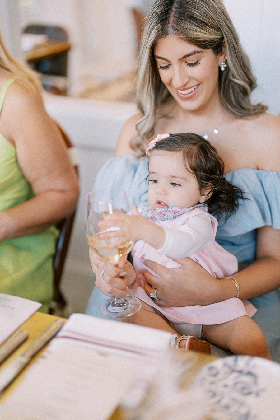 A smiling woman holding a baby on her lap at a dining table, with the baby reaching for a wine glass.