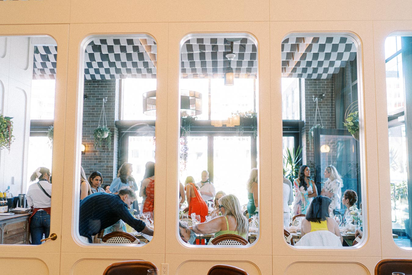 Group of people socializing in a modern restaurant with large windows, natural light, and decorative plants.