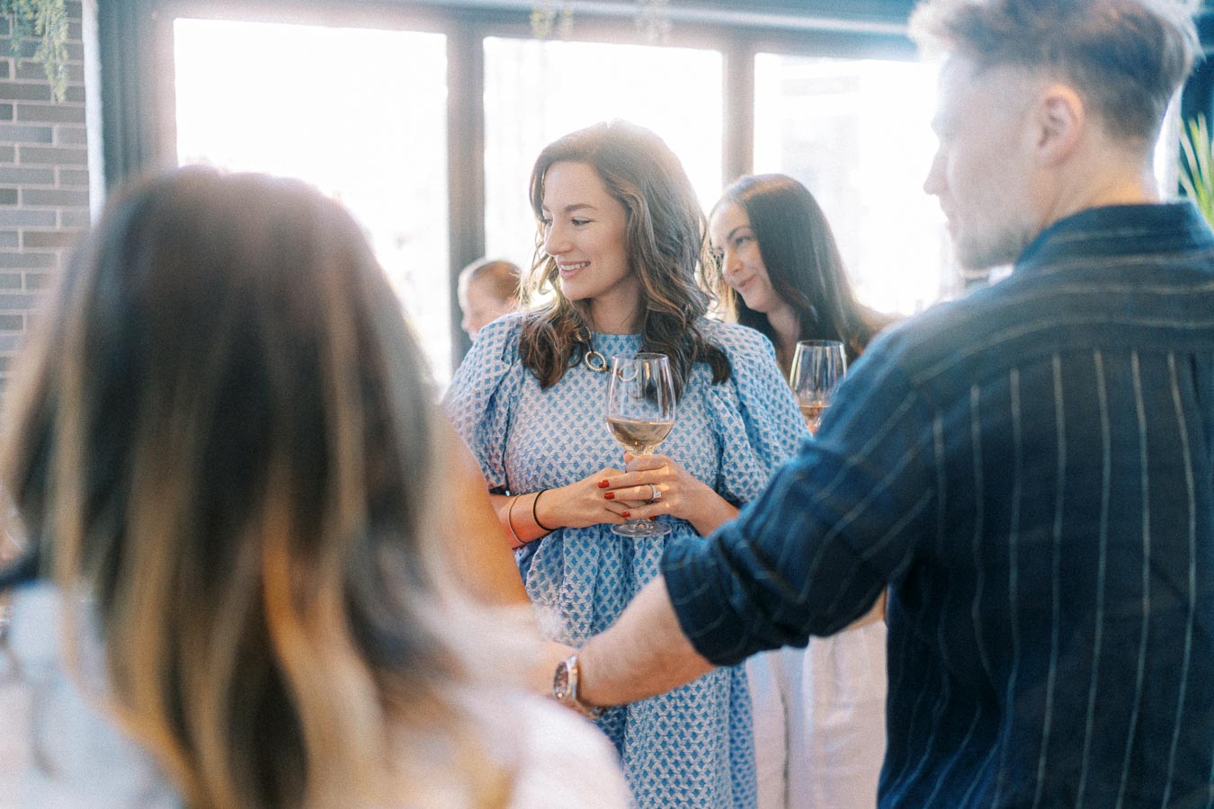 Group of people socializing at an indoor gathering, a woman smiling and holding a wine glass, wearing a blue dress, with a blurred background of friends and natural light coming from large windows.
