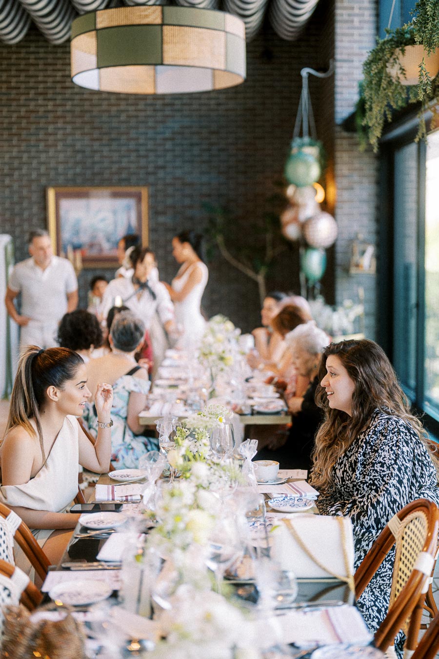 Elegant gathering in a stylish restaurant setting with guests seated at a beautifully decorated table adorned with flowers, engaged in conversation during a special event.