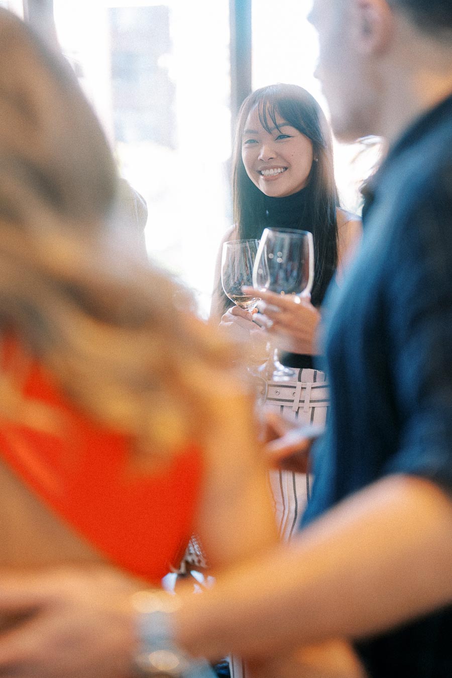 A group of people socializing at a party, where a woman is smiling and holding a wine glass, engaged in conversation in a lively setting.