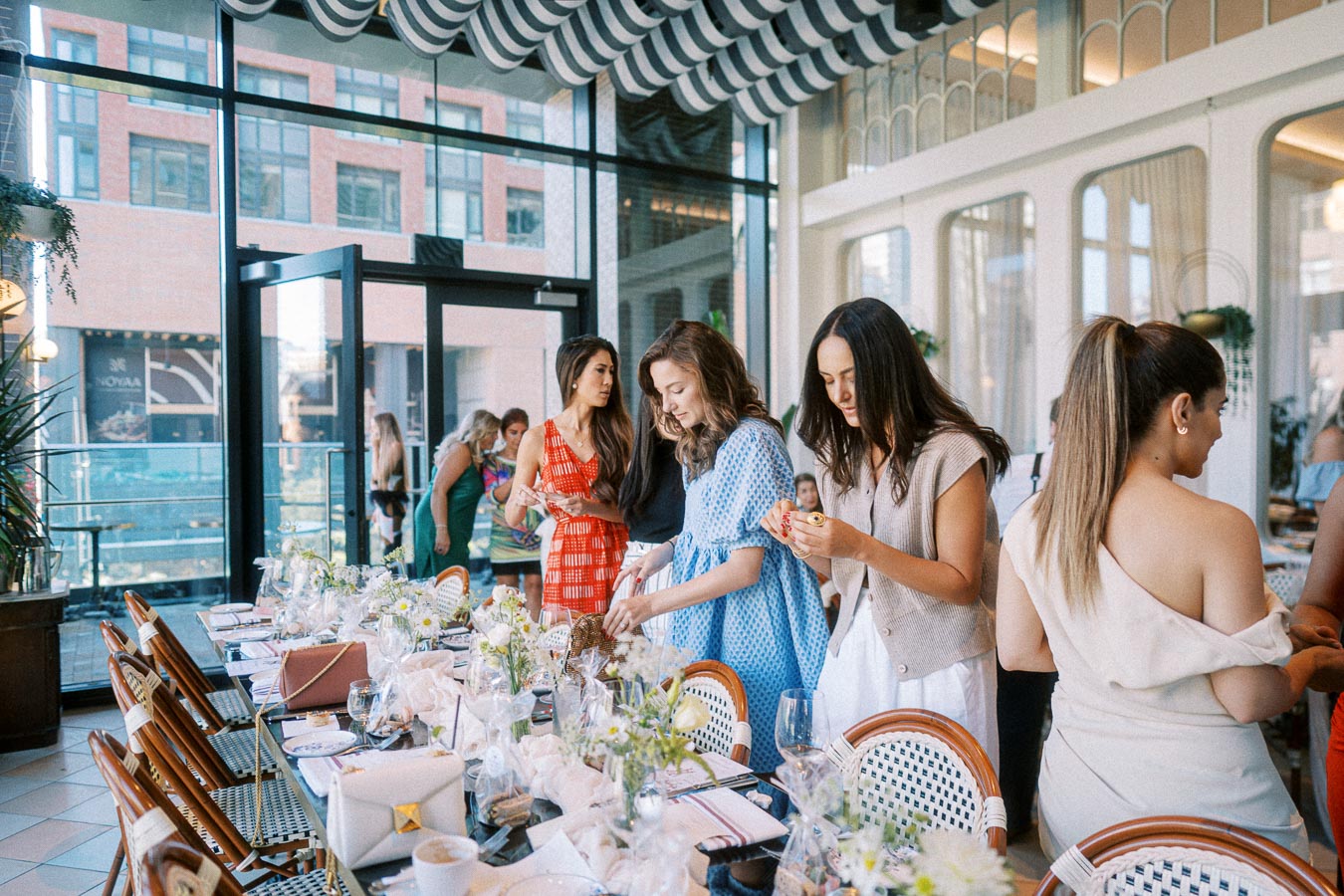 A group of women gather around a beautifully set table in a stylish restaurant with floor-to-ceiling windows, engaging in a social event. The table is adorned with flowers and elegant tableware, creating a warm and inviting atmosphere.