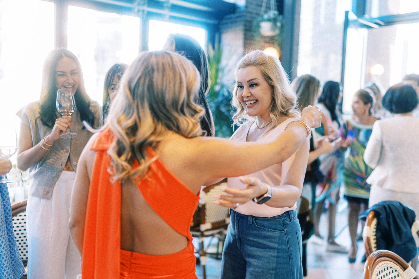 A group of women socializing and laughing at a lively party, with one woman in a bright orange dress embracing another in a casual pink top.