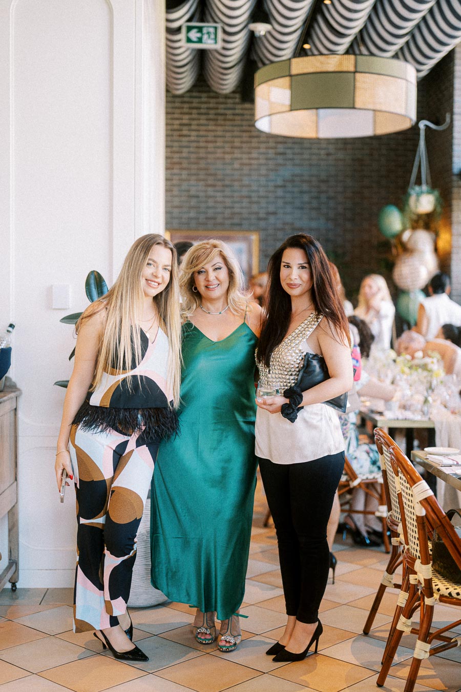 Three women smiling and posing together at a stylish indoor event, wearing colorful and elegant outfits, with a modern decor background featuring a large ceiling light and dining tables.