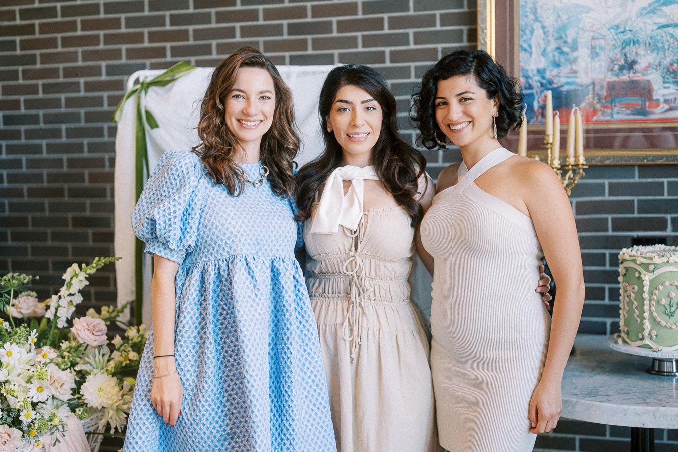 Three women smiling in elegant dresses at a celebration, posing in front of a brick wall with a floral arrangement and cake in the foreground.