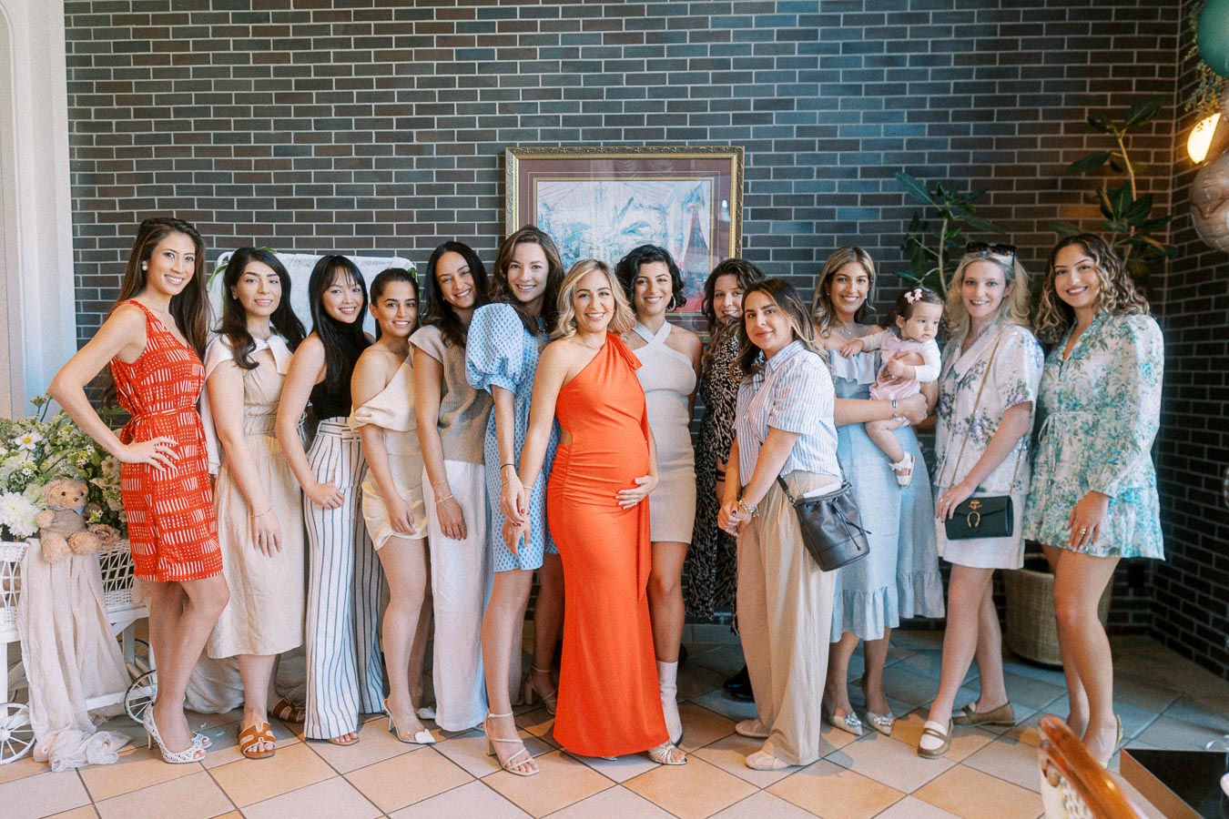 Group of women at a baby shower posing together with a brick wall backdrop, including a pregnant woman in an orange dress at the center, surrounded by friends wearing stylish outfits.