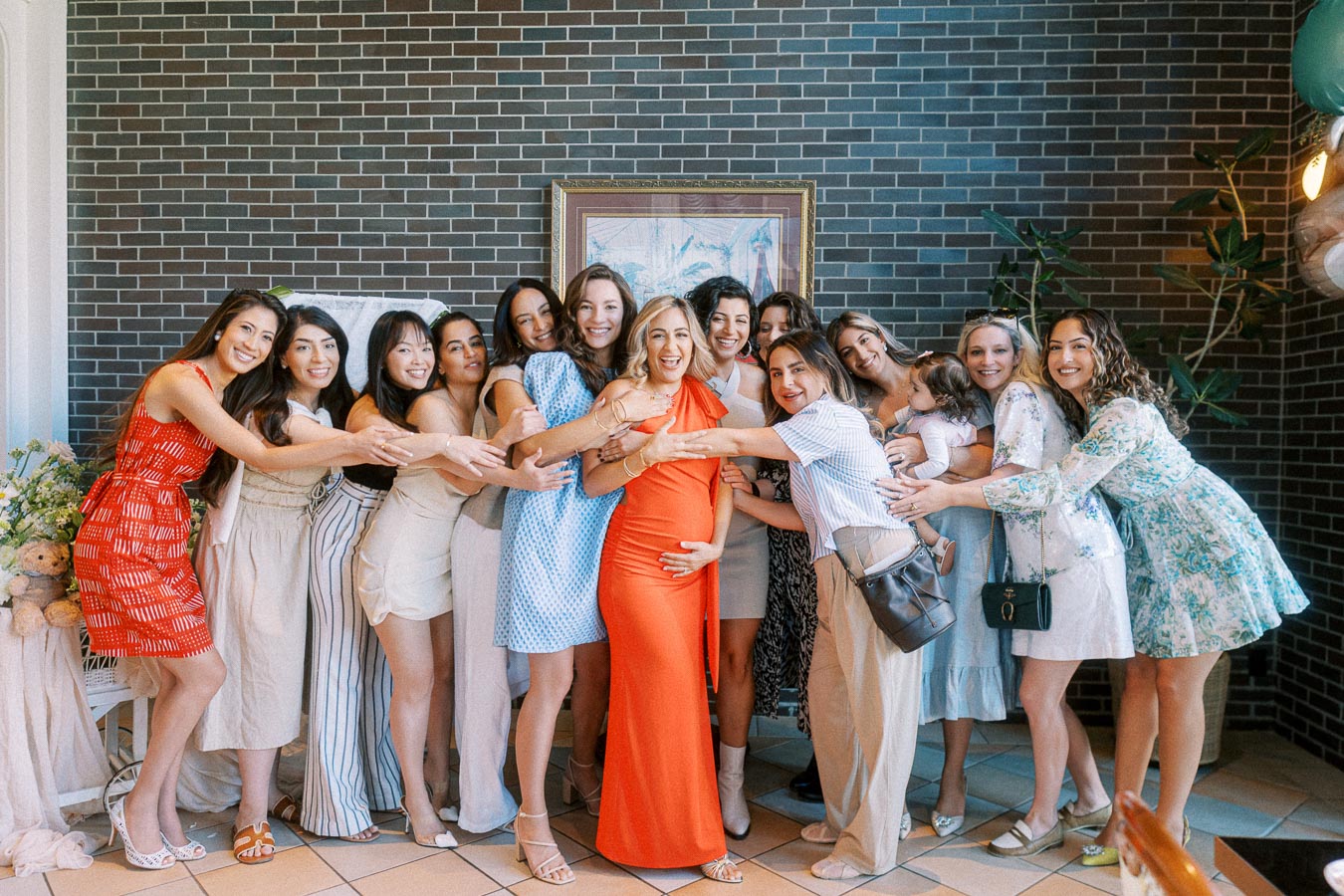 A joyful group of women celebrating a baby shower, with the expectant mother in a vibrant orange dress surrounded by friends offering loving embraces against a stylish brick wall background.
