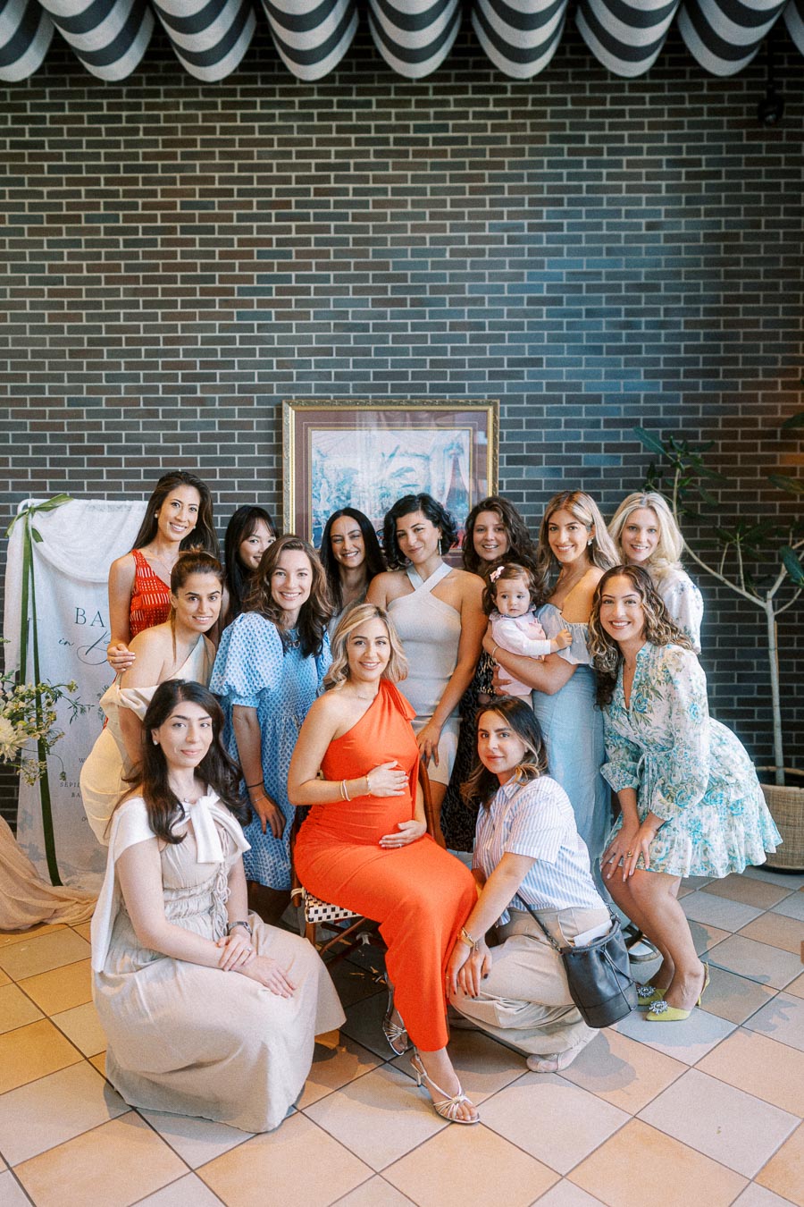 A group of women celebrating a baby shower, with the expecting mother in a vibrant orange dress sitting in the center, surrounded by friends and family in a decorated indoor space.