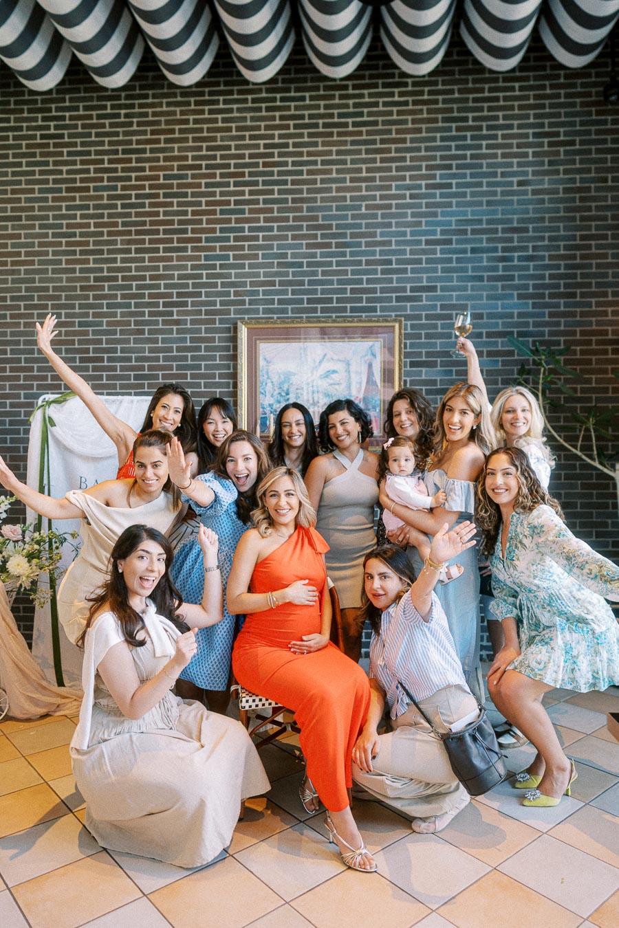 A group of joyful women celebrating a baby shower, with the expectant mother in an orange dress, surrounded by friends and family in a cheerful setting with a brick wall background.