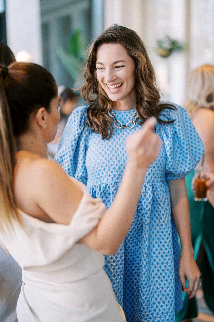 Two women engaged in conversation at a social event, one wearing a blue patterned dress and smiling warmly.