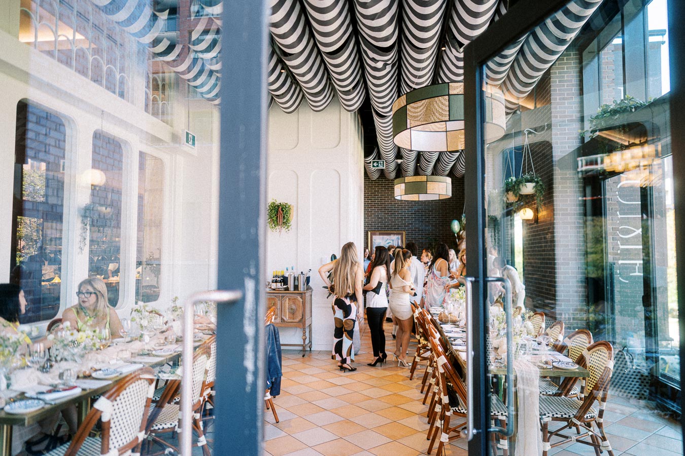 A stylish restaurant interior with guests socializing, featuring a long, elegantly set table, checkerboard floor, and unique black and white striped ceiling design.
