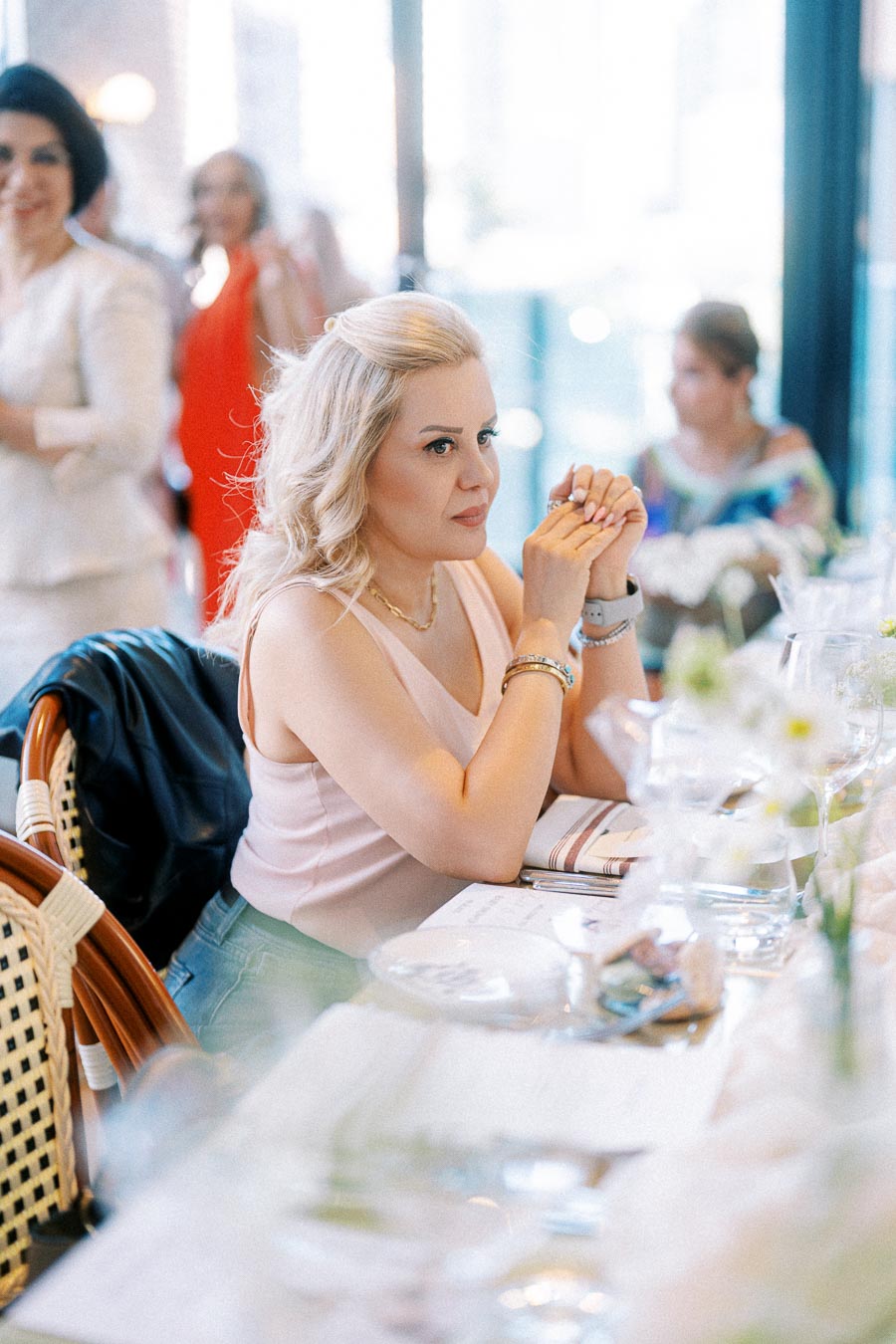 Elegant woman seated at a restaurant table, attentively listening during a social event, surrounded by blurred guests and softly lit decor.