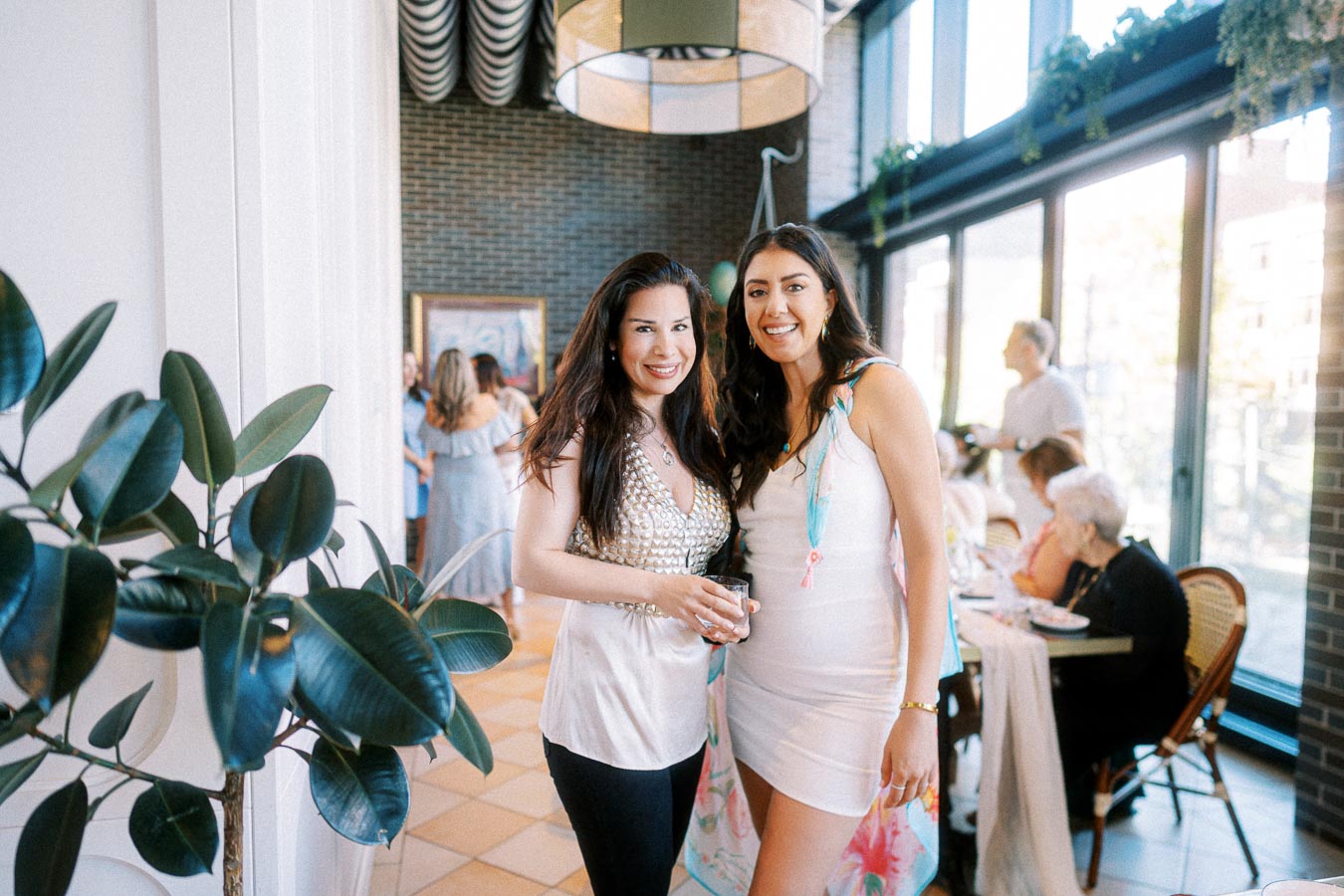 Two women smiling indoors at a social gathering, surrounded by other guests and lush green plants, in a well-lit room with brick walls and large windows.