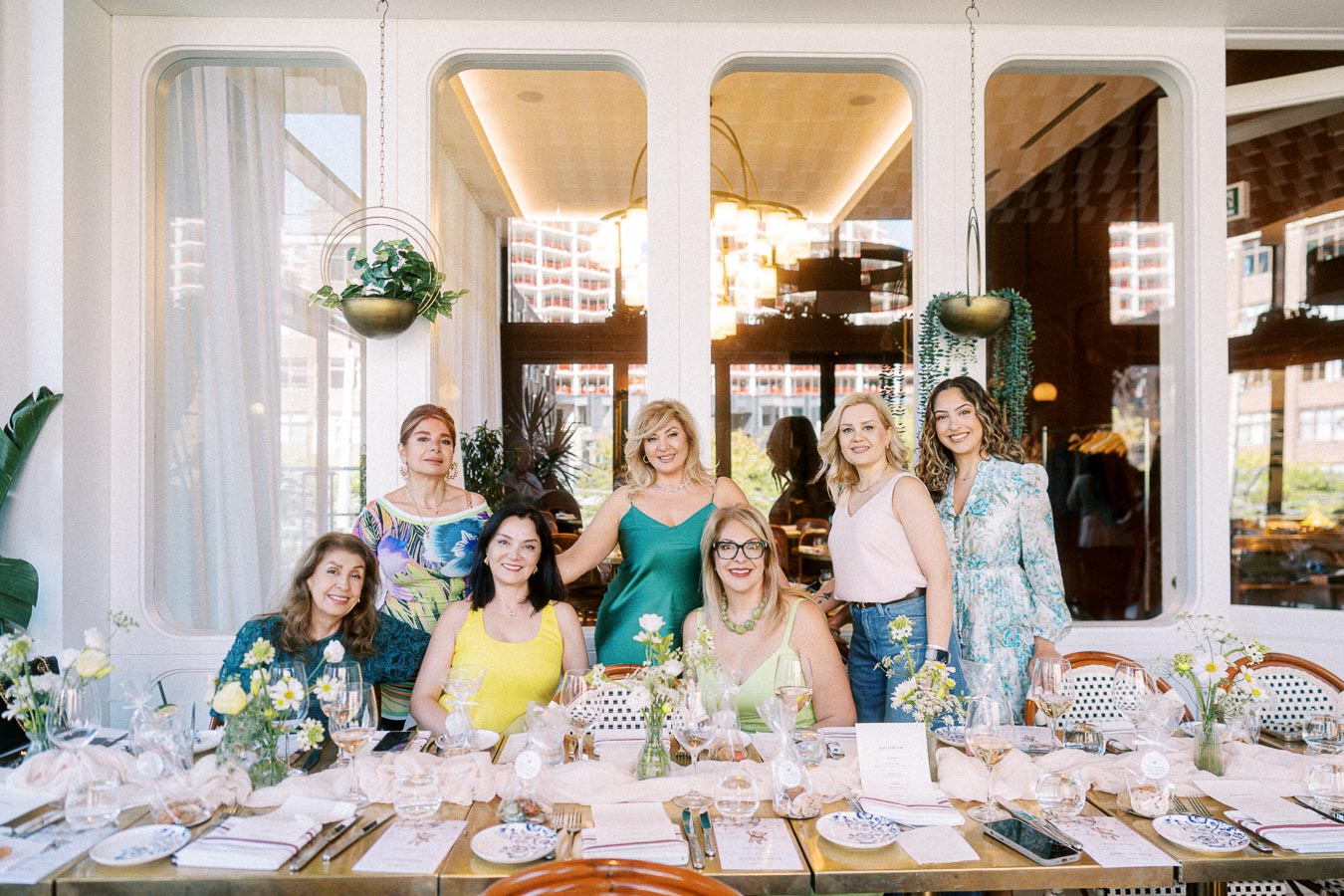 A group of women enjoying a stylish brunch together at a beautifully set dining table, surrounded by elegant decor and greenery in a well-lit, modern restaurant setting.