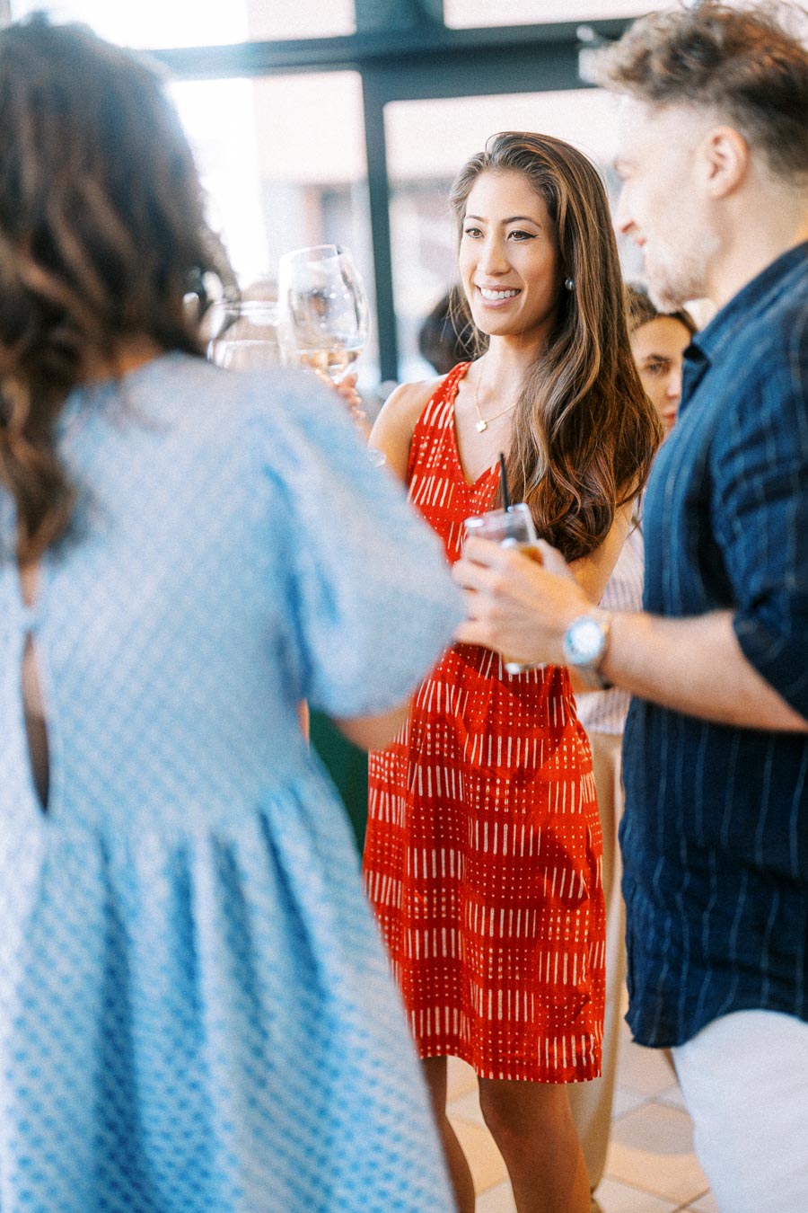 Group of friends enjoying a social gathering, smiling and toasting with drinks. A woman in a red dress stands prominently in the center, surrounded by other casually dressed attendees.