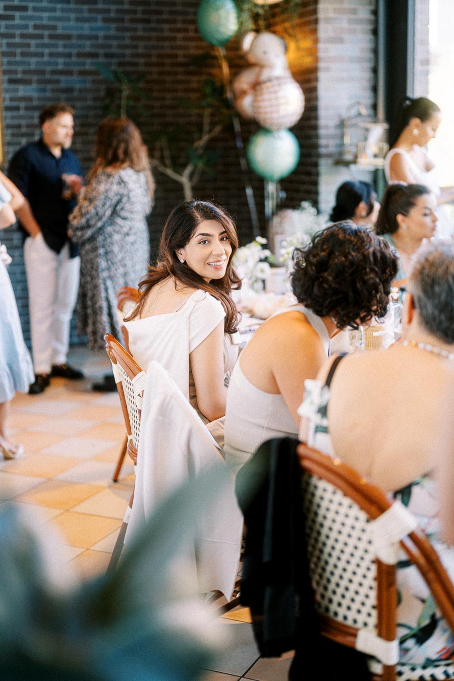 Guests enjoying a lively indoor gathering with a modern brick wall backdrop and festive decorations, highlighting a woman smiling in focus amidst a seated group.