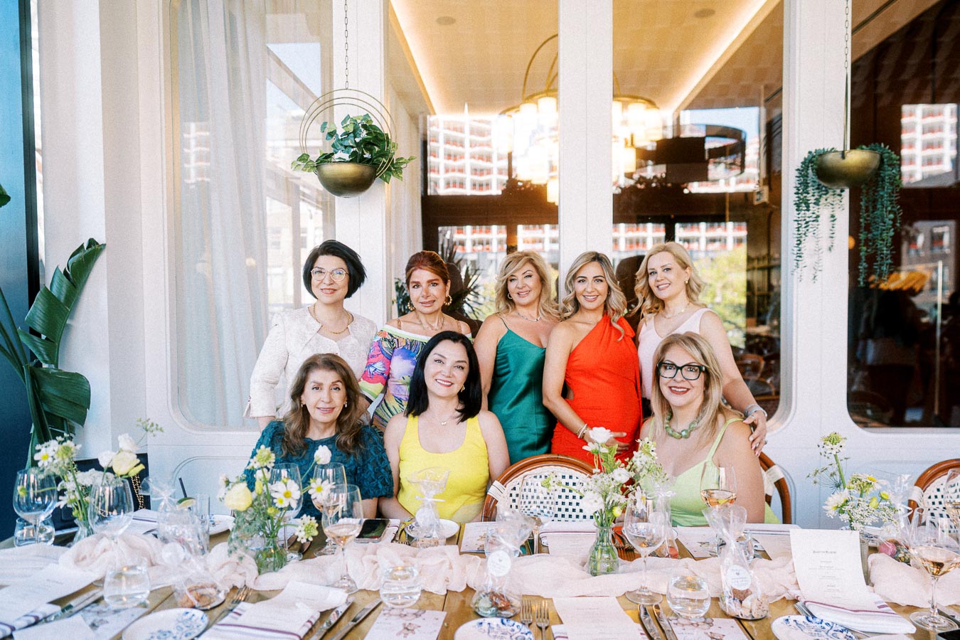 A group of elegantly dressed women smiling at a beautifully set dining table with floral arrangements in a stylish restaurant setting, featuring large windows and modern decor.