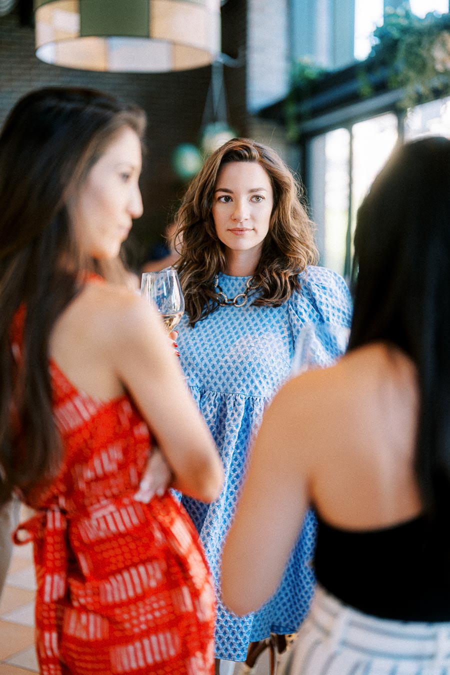 Three women at a social gathering, one in focus wearing a blue dress, engaging in conversation while holding wine glasses in a well-lit, stylish venue.