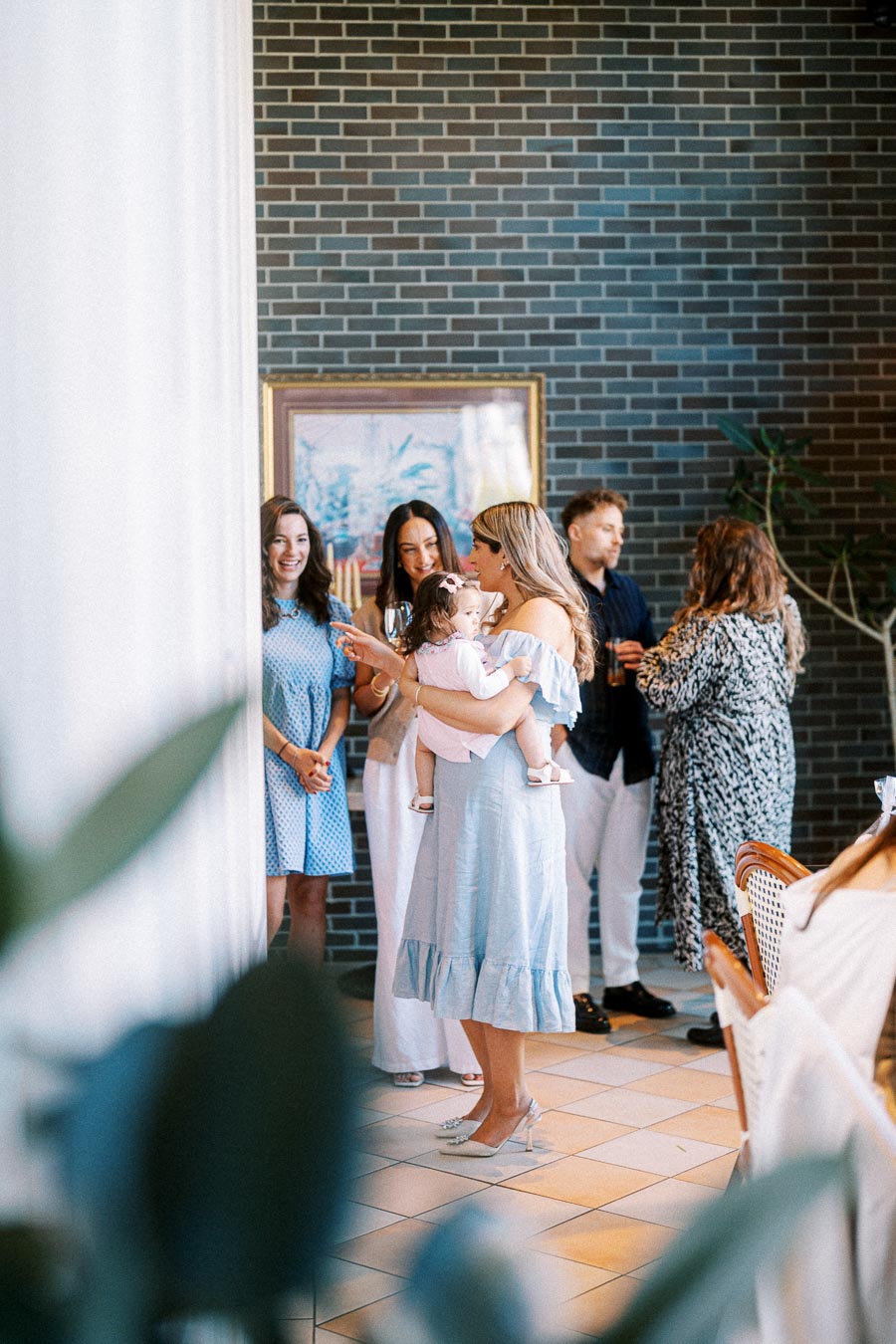 A group of people socializing indoors, with a woman holding a baby. The scene features elegant attire and a relaxed atmosphere, with a brick wall and framed artwork in the background.