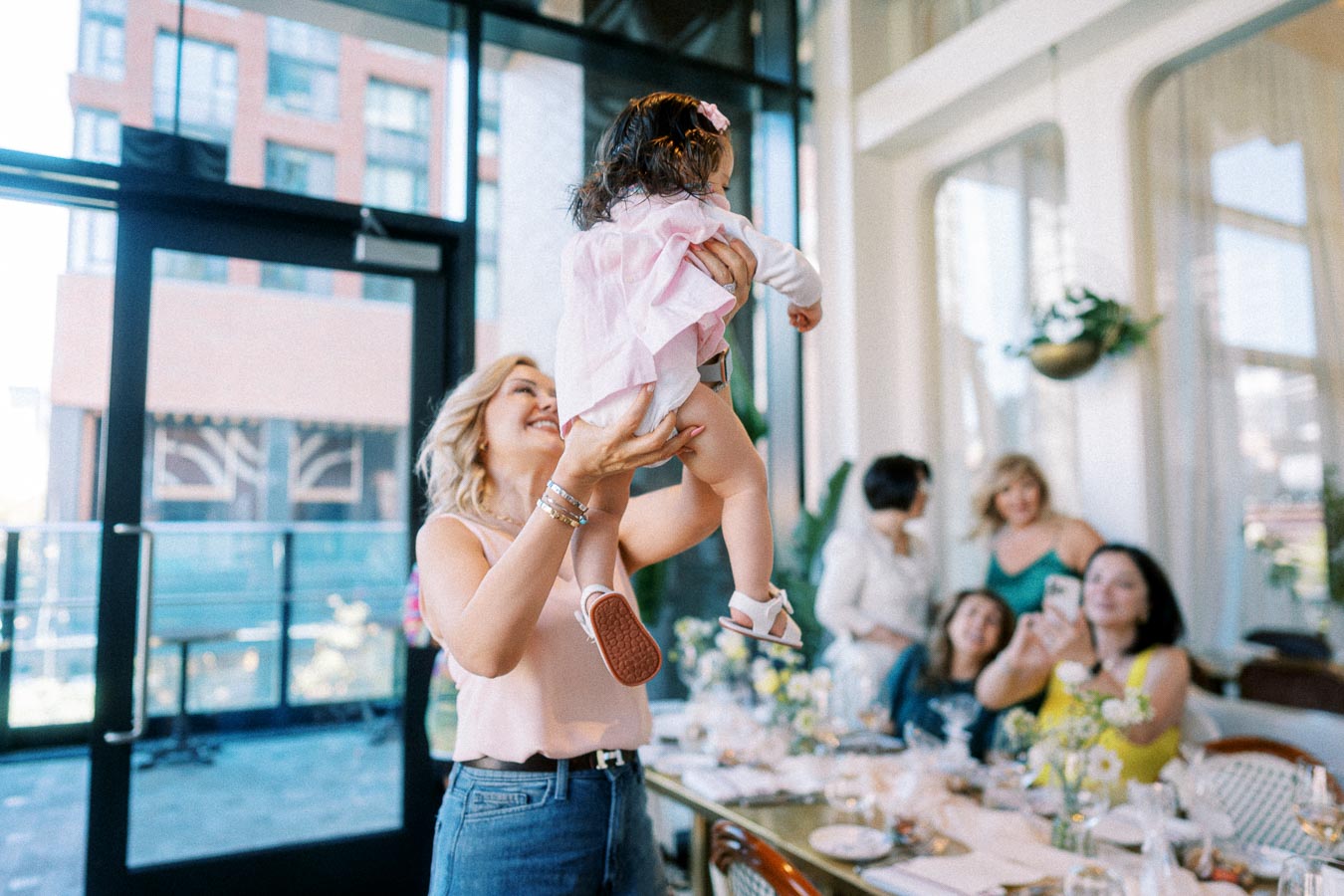 A woman joyfully lifts a toddler in a pink dress at a bright, airy gathering in a modern restaurant, surrounded by smiling friends enjoying the celebration.