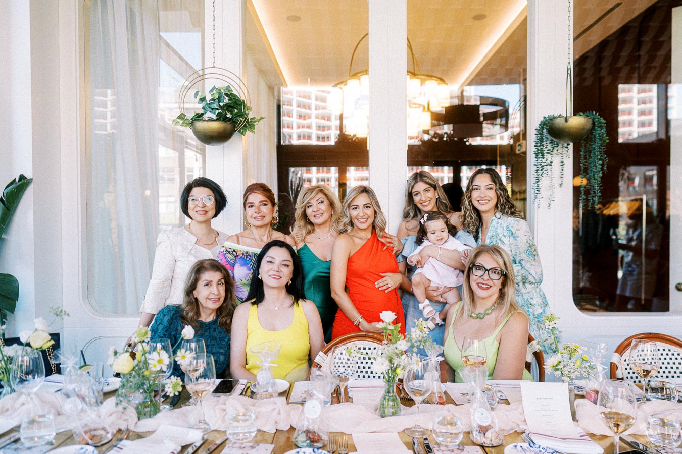A group of diverse women smiling at a beautifully set table in a stylish indoor setting, featuring elegant floral arrangements and soft lighting. The image captures a joyful gathering, highlighting a sense of community and celebration.