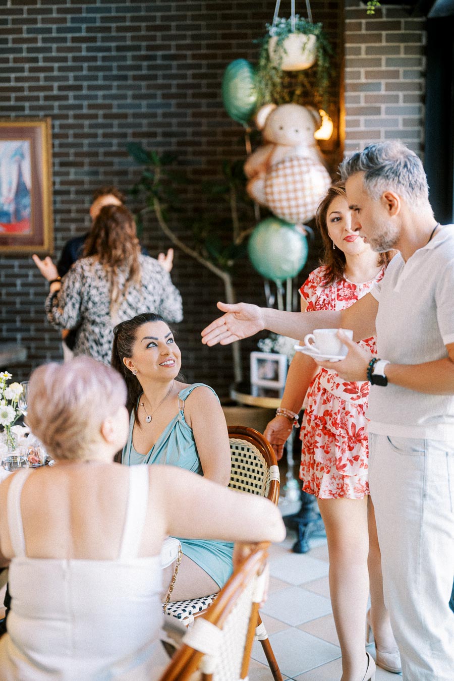 Group of people socializing at a casual indoor gathering with brick wall background and decorative balloons, featuring a man holding a cup engaging with seated guests.